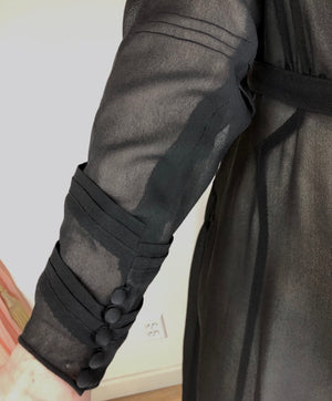 Close up of the sleeve shows sheer black fabric with two rows of 3 tucks on the lower arm and a button closure at the wrist